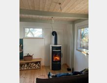 Wohnzimmer mit hellem Holzboden und Holzdecke, einem modernen schwarzen Kaminofen mit sichtbarem Feuer, einem Regal mit gestapeltem Holz sowie Fenstern, die Tageslicht einlassen. Im Vordergrund ist eine entspannte Person auf einem Sofa zu sehen.