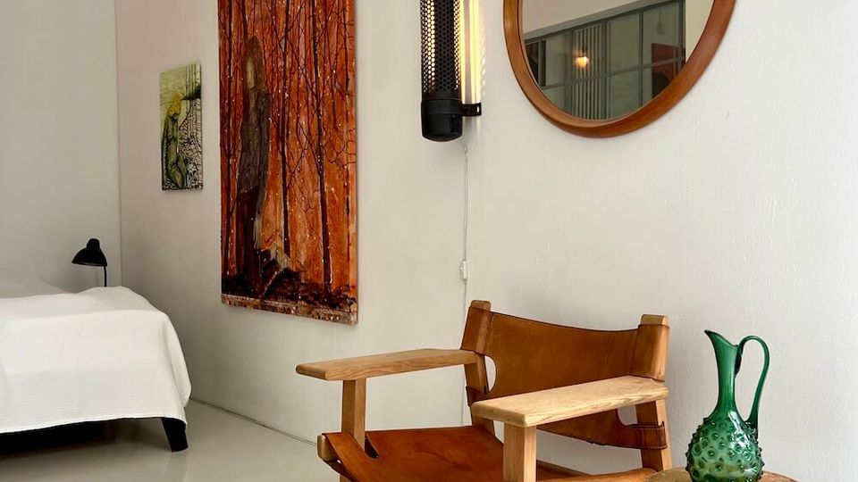 Minimalist bedroom corner with white walls and floor, featuring a low bed, a wooden lounge chair, and a small round metal side table with a green glass vase. Wall decor includes a large orange-toned painting, a smaller artwork, a vertical wall sconce, and a round wooden-framed mirror.