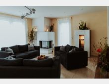 Modernes Wohnzimmer mit mehreren dunkelgrauen Sofas, einem weißen Kamin an der Wand und großen Fenstern mit transparenten Vorhängen. Dekorative Pflanzenelemente und ein beleuchteter Vitrinenschrank ergänzen den Raum.