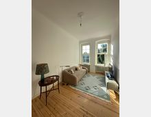 Wohnzimmer mit Holzfußboden, einem beigen Sofa und einem kleinen Beistelltisch mit einer Lampe. Große Fenster lassen Tageslicht herein, ein hellgrüner Teppich mit floralem Muster liegt auf dem Boden, und eine schlichte Deckenlampe hängt in der Raummitte.