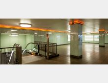 Unterirdischer Bahnhofseingang mit glänzenden Glaswänden und beleuchteten Säulen. Eine Rolltreppe führt nach unten, und die Decke ist mit runden Deckenleuchten und einer orangefarbenen Lichtleiste versehen.