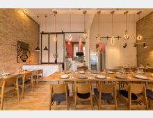 Gemütlicher Speiseraum mit Holzboden, gedeckten Holztischen und Stühlen. Die Backstein- und weiß gestrichenen Wände, moderne Hängeleuchten und eine offene Küche im Hintergrund prägen das Ambiente.