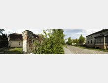 Panorama einer ländlichen Kopfsteinpflasterstraße mit alten Gebäuden, darunter ein verfallener Ziegelpfeiler links und ein Haus mit verglastem Anbau rechts. Dazwischen sind Büsche und Bäume zu sehen, die den Bereich teilweise verdecken.