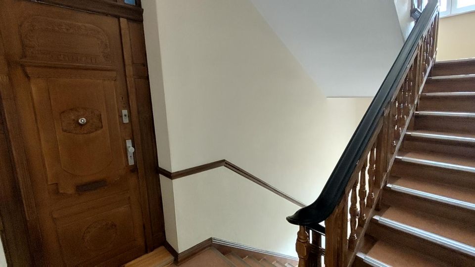 Treppenhaus in einem älteren Wohngebäude mit breiter Holztreppe und geschnitztem Holzgeländer, das zu einem Fenster im oberen Stock führt. Links befindet sich eine massive, verzierte Holztür an hell gestrichenen Wänden mit dunklen Holzleisten.