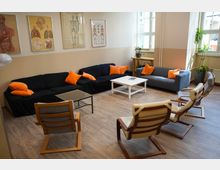 Ein gemütlicher Raum mit mehreren Sofas und Sesseln, die um einen weißen und einen gläsernen Couchtisch angeordnet sind. An den Wänden hängen medizinische Poster, und große Fenster lassen natürliches Licht herein.