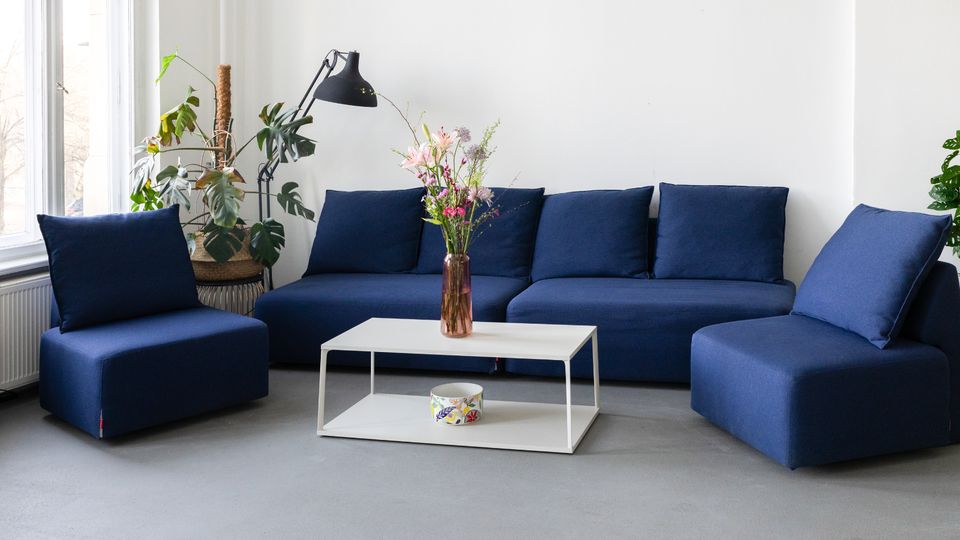 Helles Wohnzimmer mit großen Fenstern, weißen Wänden und grauem Boden, eingerichtet mit einem modularen, dunkelblauen Sofa und passendem Sessel. Davor steht ein niedriger weißer Couchtisch mit Blumen in einer Vase; daneben eine schwarze Stehlampe und mehrere Zimmerpflanzen.