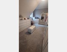 Ein Ankleidezimmer mit einem Dachschrägenfenster, mehreren weißen Kommoden, einem großen, verzierten Wandspiegel und einem kleinen weißen Sitzhocker mit Stauraum in der Mitte. Der Raum ist mit hellgrauem Teppichboden ausgelegt und wirkt ordentlich eingerichtet.