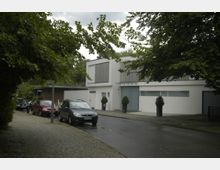 Moderne Wohngegend mit einem weißen, kubischen Haus mit großen Fenstern und flachem Dach. Im Vordergrund ist eine gepflasterte Straße mit parkenden Autos und dichter Baumbegrünung zu sehen.