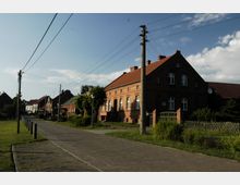 Straßenszene in einem Dorf mit gepflastertem Weg, umgeben von rotbraunen Backsteinhäusern mit roten Dächern. Im Vordergrund befindet sich ein Strommast mit gespannten Kabeln, und im Hintergrund sind weitere Häuser und grüne Gärten zu sehen.