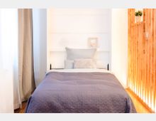 Helles Schlafzimmer mit einem Einzelbett, das eine graue gesteppte Decke und mehrere helle Kissen hat. Links ist ein Fenster mit hellen Vorhängen, rechts eine Holzvertäfelung mit Beleuchtung und ein kleiner dekorativer Blumentopf.