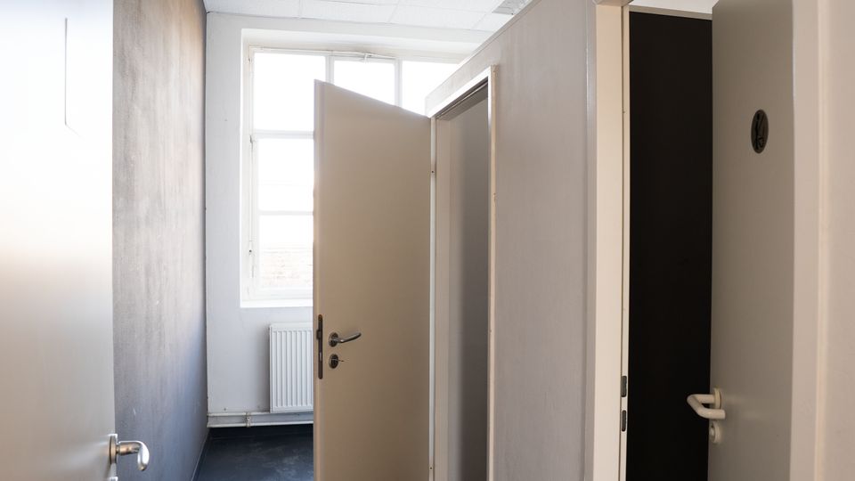 Schmaler Innenflur mit hellen Wänden und abgehängter Rasterdecke, am Ende ein großes Fenster über einem Heizkörper. Mehrere Türen stehen offen und führen in angrenzende, teils dunkle Räume.