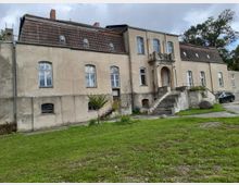 Großes, zweistöckiges historisches Gebäude mit hell-beigem Putz, symmetrischen Fenstern und einem kleinen Balkon über einem dekorativen Eingangsportal mit steinernen Treppen. Im Vordergrund eine gepflegte Rasenfläche, im Hintergrund Bäume und bewölkter Himmel.