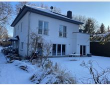 Zweistöckiges, weiß verputztes Wohnhaus mit flachem Walmdach, Schornstein und großen Fensterfronten sowie Schiebetüren zur Terrasse. Im Vordergrund liegt ein verschneiter Garten mit Sträuchern, einem Tisch auf der Terrasse und kahlen Bäumen vor blauem Winterhimmel.
