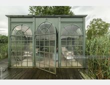 Grünes, verglastes Gartenhäuschen mit drei großen, bogenförmigen Fenstern, von denen eines als Tür geöffnet ist. Das Häuschen steht auf einer Holzplattform und ist von hohen Gräsern und Bäumen umgeben.