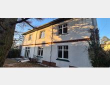 Zweistöckiges Wohnhaus mit weiß verputzter Fassade, roten Klinkerakzenten und grünen Fensterrahmen, umgeben von Bäumen und Sträuchern. Die Aufnahme zeigt die beleuchtete Seite des Gebäudes bei Tageslicht.