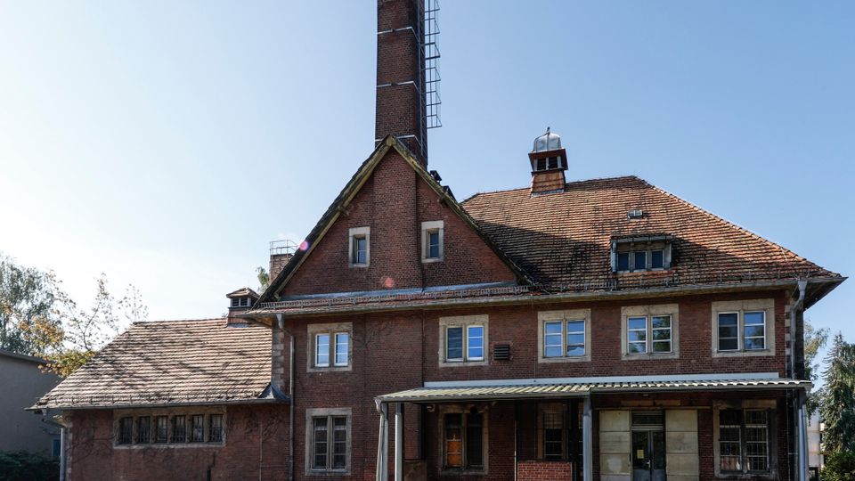 Backstein-Industriegebäude mit steilem Ziegeldach und hoher Schornsteinanlage, vorne ein überdachter Eingangsbereich mit Metallstützen. Mehrere rechteckige Fenster und kleine Dachaufbauten sind sichtbar, davor ein gepflasterter Bereich mit Geländer unter klarem Himmel.