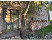 Außenansicht eines alten Backsteingebäudes mit verwitterter, grün gestrichener Holztür und kleiner Betontreppe. Daneben stehen ein Baum und ein hoher Stapel Ziegelsteine an der Wand, umgeben von Gras und niedrigem Bewuchs.