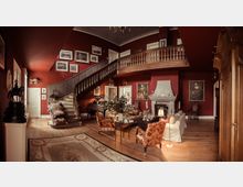 Wohnzimmer im klassischen Stil mit roten Wänden, einer hölzernen Wendeltreppe und einem Kamin. Der Raum ist mit antiken Möbeln, Gemälden, einem Teppich und dekorativen Elementen wie Blumen und Kerzen eingerichtet.