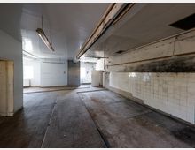 Leerer, heruntergekommener Industrie- oder Küchenraum mit gefliestem Boden, weißen Wandfliesen und deutlichen Ruß- und Schmutzspuren an der rechten Wand. An der Decke hängen lange Leuchtstofflampen und es verlaufen offene Leitungen; im Hintergrund sind Fenster und eine Tür zu sehen.