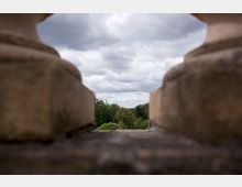 Blick durch eine steinerne Balustrade auf eine bewaldete Landschaft mit einem grünen Kuppeldach eines Gebäudes im Hintergrund unter einem bewölkten Himmel.