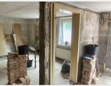 Ein renovierungsbedürftiger Innenraum mit unverputzten Wänden, einem offenen Türrahmen und sichtbarem Mauerwerk. Im Raum stehen gestapelte Ziegelsteine, Eimer mit Werkzeugen und Baumaterialien, sowie ein Fenster, das Tageslicht hereinlässt.