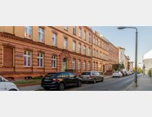 Straßenszene mit historischen Wohngebäuden aus Ziegelsteinen, die durch gelbe und rote Muster verziert sind. Parkende Autos säumen die Straße, und eine Straßenlaterne steht auf dem Gehweg.