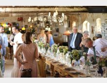 Innenraum einer rustikalen Veranstaltungslocation mit langen Holztischen, die mit Blumensträußen, Glasgefäßen und Geschirr gedeckt sind. Menschen in festlicher Kleidung unterhalten sich, während im Hintergrund nostalgische Dekorationen und Lampen zu sehen sind.