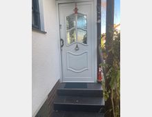 Weiße Haustür mit dekorativem Glasfenstereinsatz und einem roten Anhänger darüber, erreichbar über eine kleine Treppe mit dunklen Fliesen. Links ist eine verputzte Wand und rechts eine Holzstütze mit Pflanzen und weihnachtlicher Dekoration.