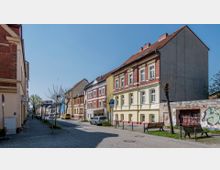 Straßenszene mit historischen Wohnhäusern, teils in gelben und roten Fassadenfarben, entlang einer gepflasterten Straße mit Gehweg. Im Vordergrund stehen Parkbänke und junge, beschnittene Bäume, während am Straßenrand Autos und ein Verkehrsschild sichtbar sind.