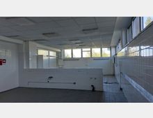 Ein leerer, gefliester Raum mit weißen Wänden, großen Fenstern im oberen Bereich, einer abgehängten Decke mit integrierten Leuchtkörpern und grauem Fliesenboden. Im Vordergrund sind Wasseranschlüsse und ein Waschbecken sichtbar, im Hintergrund ein freier Blick auf die Fenster, die Tageslicht hereinlassen.