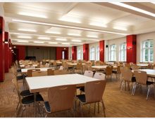 Ein großer, heller Speisesaal mit weißer Decke, roten Wandelementen und mehreren Reihen aus Holztischen und Stühlen auf einem Parkettboden. Große Fenster an der rechten Wand lassen natürliches Licht herein.