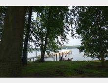 Ein See mit einem Steg und mehreren gelben und roten Pfeilern im Hintergrund, umgeben von Bäumen. Im Vordergrund sitzen Personen in einem Kanu am Ufer auf einer grasbewachsenen Fläche.