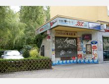 Ecke eines kleinen Ladens mit geschlossener, grafittibeschmierter Metallrolltür und der Aufschrift "Tabakwaren". Die Fassade trägt Plakate und Werbeschilder für Zeitungen und Tabakmarken, daneben ein alter Zigarettenautomat; im Hintergrund sind eine Hecke, Bäume und ein geparktes Auto zu sehen.