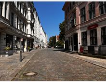 Pflastersteinstraße in einer Altstadt mit historischen Gebäuden auf beiden Seiten, darunter ein rosafarbenes Gebäude mit der Aufschrift "Museum". Im Hintergrund sind Bäume und weitere Gebäude sichtbar, bei strahlend blauem Himmel.