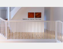 Innenansicht einer Galerie mit weiß gestrichenen Wänden und einem hölzernen Boden. Im Mittelpunkt befindet sich ein Gittergeländer, dahinter hängen zwei abstrakte Kunstwerke in warmen Orangetönen an der Wand, darüber der Schriftzug „Galerie am kleinen Wannsee“.