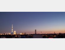 Blick auf die Skyline von Berlin bei Abenddämmerung, mit dem beleuchteten Fernsehturm als markantem Wahrzeichen links und flachen Dächern im Vordergrund. Der Himmel zeigt einen Farbverlauf von Blau zu Orange.