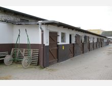 Einstöckiger, länglicher Stallbau mit weiß verputzten Wänden, braunen Türen und Fenstern. Der gepflasterte Hof davor zeigt landwirtschaftliches Gerät und Holzpaletten an der Wand.