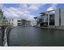 Moderne Regierungsgebäude mit großer Glasfassade entlang eines künstlichen Wasserkanals, umgeben von Gehwegen und Bäumen. Der Himmel ist teils bewölkt.