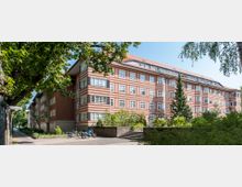 Mehrstöckiges Wohngebäude mit rot-beigem Streifenmuster und zahlreichen weißen Fenstern, umgeben von Bäumen und Grünflächen. Im Vordergrund ein Weg mit Treppenstufen, Fahrrädern und einem Straßenschild mit "Hortensienplatz".