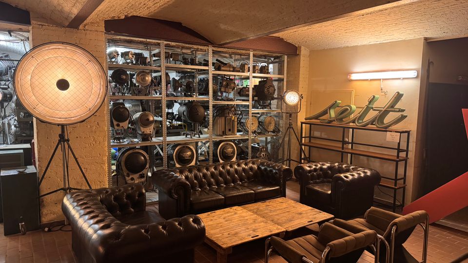 Industrie-inspirierter Raum mit freiliegenden Ziegelwänden, braunen Ledersofas im Chesterfield-Stil und einem niedrigen Holztisch. Im Hintergrund stehen Regale mit vintage Bühnen- und Filmbeleuchtung sowie ein leuchtendes Schriftzeichen "seit".
