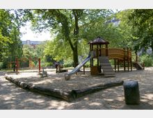 Ein Spielplatz mit sandigem Boden, einer hölzernen Kletter- und Rutschstruktur sowie Schaukeln im Hintergrund, umgeben von hohen Bäumen und einigen Wohngebäuden im Hintergrund.