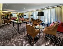 Eleganter Lounge-Bereich mit gepolsterten Sesseln und Sofas in warmen Farben, Glastischen, großen Stehlampen und regalmäßig angeordneten Büchern im Hintergrund. Bodentiefe Vorhänge und ein gemusterter Teppich prägen die stilvolle Atmosphäre.