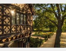 Historisches Fachwerkgebäude mit detailliertem, geometrischem Muster an der Fassade, umgeben von einem gepflegten Garten mit Blumenbeeten, Hecken und einem großen Baum im Vordergrund. Die Szenerie ist in warmes Sonnenlicht getaucht.