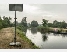 Ein schmaler Fluss fließt durch eine bewaldete Landschaft mit Schilf am Ufer, daneben ein Sandweg. Im Vordergrund steht ein Straßenschild, rechts ist eine schmale Brücke mit einem Warnschild zu sehen.