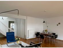 Wohnraum mit modernem Design: Ein Sitzbereich mit einem blauen Sessel und einem Glastisch auf einem weißen Teppich, daneben ein Esstisch aus Holz mit sechs schwarzen Stühlen. Im Hintergrund eine weiße Wendeltreppe, weiße Vorhänge und eine teilweise offene Küche.