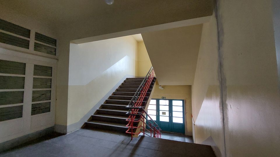 Treppenhaus mit einfachen, gelb-beigen Wänden, einer Treppe mit dunklen Stufen und einem roten Geländer. Im Hintergrund befinden sich eine Glastür mit grünem Rahmen und ein Notausgangsschild darüber.