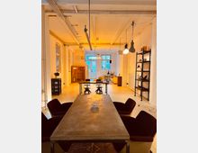 Großer, offener Raum mit industriellem Stil, weißen Wänden und freiliegenden Deckenbalken. Im Vordergrund steht ein langer Tisch mit roten Polsterstühlen, im Hintergrund befinden sich weitere Tische, Regale und große Fenster, die den Raum beleuchten.