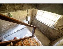 Blick in einen alten Keller- oder Treppenhausbereich mit roh verputzten, teils freiliegenden Ziegelwänden und einer Holztreppe mit gemusterten Fliesenstufen. Rechts fällt Tageslicht durch ein vergittertes Fenster, an der Wand hängen ein kleiner Elektroverteiler und Leitungen.
