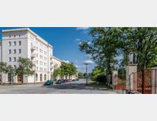 Straßenansicht mit einem mehrstöckigen Wohngebäude im klassischen Stil auf der linken Seite, umgeben von Bäumen und geparkten Autos. Rechts befindet sich ein rotes schmiedeeisernes Tor mit steinernen Pfosten, das zu einem begrünten Bereich führt.