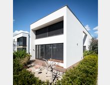 Modernes Wohnhaus mit weißer Fassade und großen Fenstern mit schwarzen Lamellenrollos. Im Vordergrund befindet sich ein kleiner Garten mit Kiesfläche, einem kahlen Baum und Sitzmöbeln; umgeben von Buschhecken.